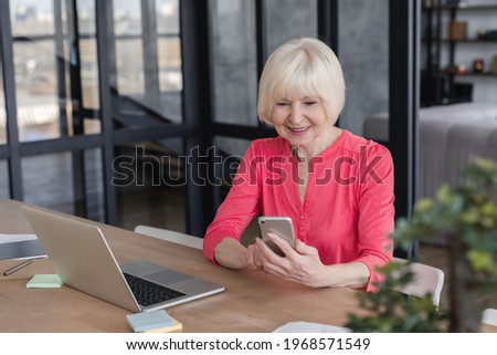 Smiling senior old elderly businesswoman grandmother using smart phone for remote work, talking with clients, loading feed, communicating with colleagues and grandchildren at home office
