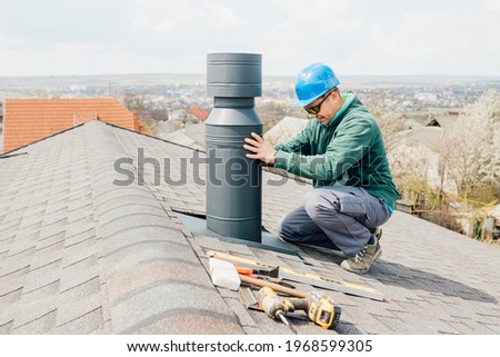 male worker with blue helmet installs an iron chimney. worker with sunglasses repairing chimney on roof. Royalty-Free Stock Photo #1968599305