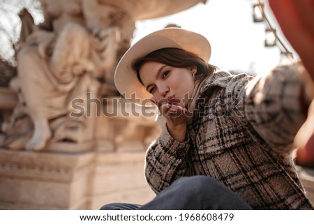 Stylish woman takes selfie and blows kiss on street. Beautiful short-haired girl in beige hat, checkered coat and jeans sits near fountain.