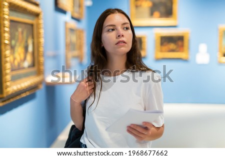 Focused young girl visitor holds a booklet with an exhibition program while admiring the paintings in the museum Royalty-Free Stock Photo #1968677662