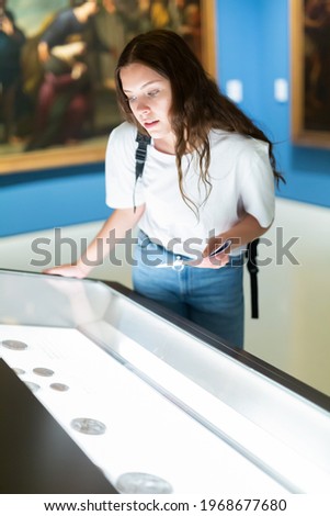 Attentive girl visitor at the museum looks at the exposition behind a glass display case, holding an information ..booklet Royalty-Free Stock Photo #1968677680