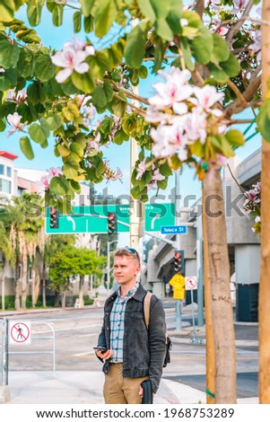 A young man in a denim jacket uses a smartphone while standing on a Los Angeles street next to a pink flowering tree