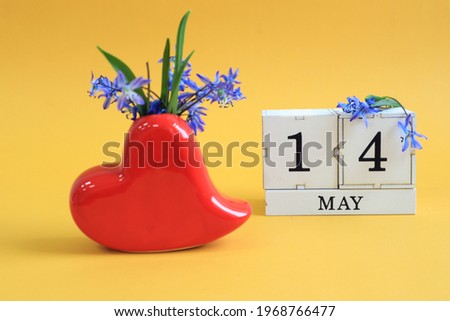Calendar for May 14: a bouquet in a heart-shaped vase with blue flowers and the numbers 14 on cubes, the name of the month of May in English, yellow background