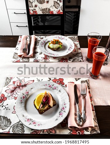 festive kitchen table setting and food with drinks and beautiful red-green tablecloths with flower patterns. Shrimp sandwich, herring sandwich, tomato juice, lemon, herbs,, a vase of flowers. Tableclo