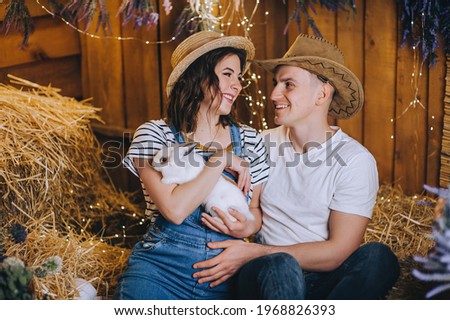 A loving man cowboy and a pregnant woman in denim overalls and a straw hat are sitting in the hay with a white rabbit in their arms, smiling and hugging in the countryside. Photography, concept.