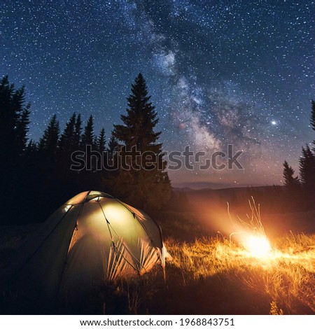 Tourist camp near forest in warm summer night. Illuminated tent and bright campfire burning under beautiful night sky full of stars and shiny milky way. Concept of camping, tourism, outdoor lifestyle.