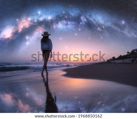 Arched Milky Way and young woman in hat on sandy beach against starry sky reflected in water at night. Landscape. Summer travel. Girl on the tropical sea coast and bright Milky Way arch, stars. Space	