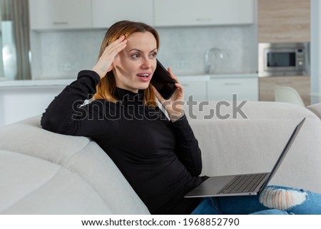 Beautiful young girl talking loudly on the phone working behind a laptop, sitting on the couch in her house. High quality photo