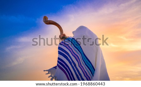 Blowing the shofar for the Feast of Trumpets - Jewish man in a traditional tallit prayer shawl blowing the ram's horn against beautiful sunset sky Royalty-Free Stock Photo #1968860443