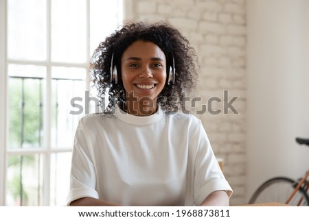Happy young 25s african american businesswoman wearing wireless headphones looking at camera, holding video call distant conversation, negotiating working issues with colleagues by virtual event. Royalty-Free Stock Photo #1968873811