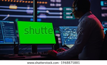 Professional IT Technical Support Specialist and Software Developer Working on Computer with Green Screen Mock Up Display in Monitoring Control Room with Digital Screens. Employee Uses on Headphones.