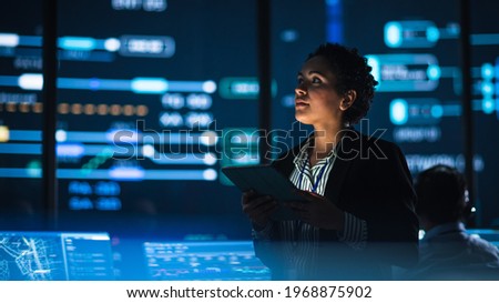 Young Multiethnic Female Government Employee Uses Tablet Computer in System Control Monitoring Center. In the Background Her Coworkers at Their Workspaces with Many Displays Showing Technical Data. Royalty-Free Stock Photo #1968875902