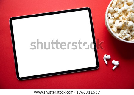 Tablet with empty white screen with wireless earphones and bowl of popcorn next to it. Mockup for video or movie screenshot. Royalty-Free Stock Photo #1968911539