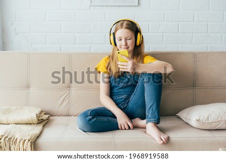 Teenage girl sitting on the couch plays the phone and listens with wireless headphones. Summer school holidays. Time for mobile games. Video call, online communication, social networks, friends. Royalty-Free Stock Photo #1968919588