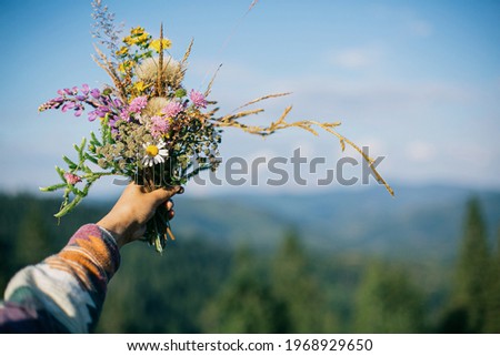 Woman traveler holding bouquet of wildflowers on background of mountain hills and sky. Hiking in mountains and gathering colorful beautiful flowers. Wanderlust and travel concept. Royalty-Free Stock Photo #1968929650