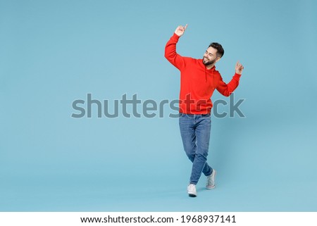 Full length young caucasian happy bearded overjoyed relaxed excited man 20s in casual red orange hoodie stand with raised up hands index fingers enjoying isolated on blue background studio portrait