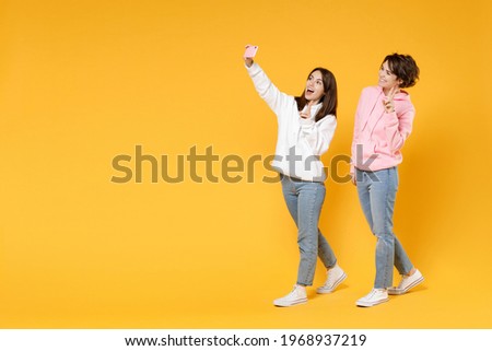 Full length of excited two young women friends 20s wearing casual white pink hoodies doing selfie shot on mobile phone showing victory sign isolated on bright yellow color background studio portrait