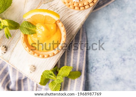 Shortbread tartlet filled with lemon curd, mint and lemon slices and mini meringue on wooden board, blue concrete background. Top view. Selective focus.