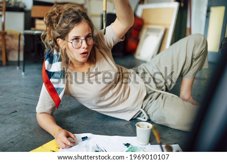 Horizontal image of a female artist sitting on the floor in the art studio and painting on paper with a brush. A beautiful woman painter with glasses painting with watercolors in the workshop.