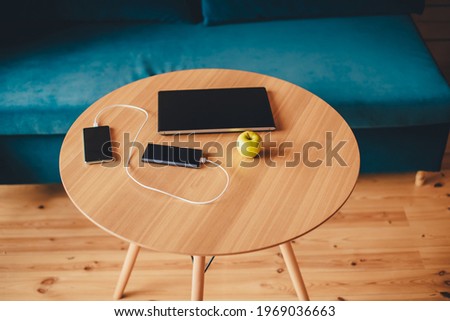 Sunlight illuminates living room workplace home office interior for comfort productive work. on a wooden table a laptop, a mobile phone is charged from a power bank and a green apple. 