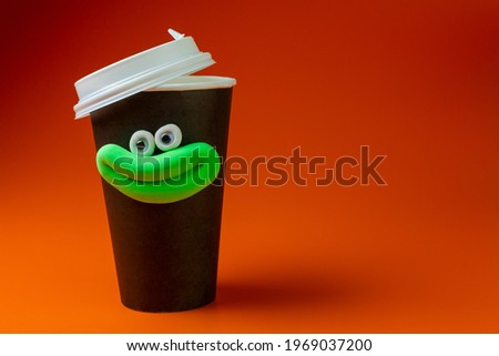 Coffee cup with eyes and smiling mouth on orange background. Cartoon plasticine parts of face on things. For advertisement coffee to go. Space for text