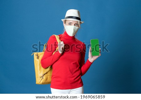 Girl in mask, hat, with backpack shows green screen of the phone. Smartphone application advertising concept.Topic of new travel rules during a pandemic. Studio blue background. Red, white, blue color