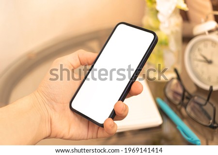 Man hold mobile phone mockup screen on background of home desk with alarm clock, glasses and homeplant, home interior concept 