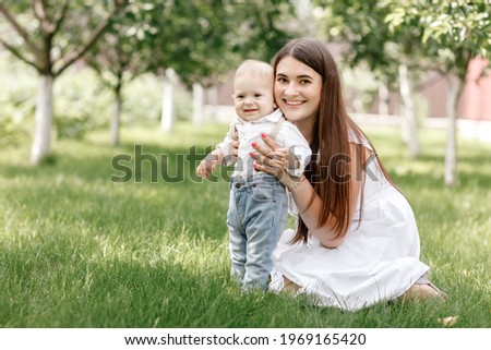 Happy beautiful woman, young mother playing with her adorable baby son, cute little boy, enjoying together a sunny warm day playing on the lawn in a summer garden.