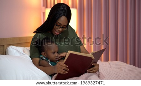 african american mother reading a book to her little daughter in bed before going to sleep at home. black mom story telling to girl in bedroom Royalty-Free Stock Photo #1969327471