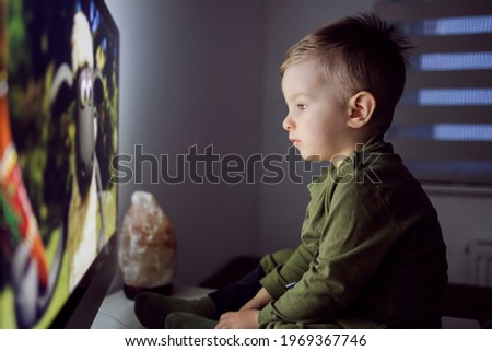 Modern parenting. A baby boy is sitting right in front of the TV and staring at a cartoon screen. Entertaining a child before going to bed at night in the age of advanced technology
