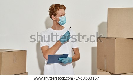 Picture of a young deliveryman keeps track of parcels, young deliveryman counts cardboard boxes with pen, courier holds clipboard in hands, watches aside