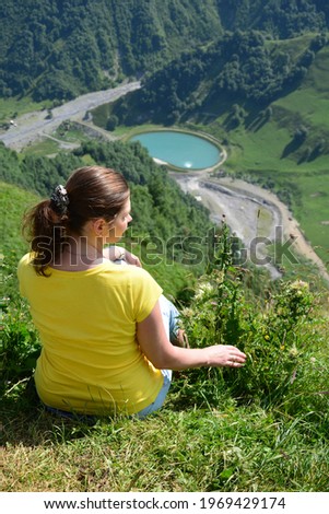 Beautiful young woman model in  yellow T-shirt sits on a top of green mountain meadow and admires the turquoise mountain blue lake and the surrounding stunning nature. Summer in the Caucasus Mountains