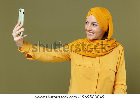Young smiling happy friendly arabian asian muslim woman 20s in abaya hijab yellow clothes doing selfie shot on mobile phone isolated on olive green background studio People uae islam religious concept