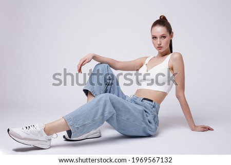 High fashion photo of a beautiful elegant young woman in a pretty white top and sneakers, blue denim jeans posing over white, soft gray background. Studio Shot. The model is sitting Royalty-Free Stock Photo #1969567132