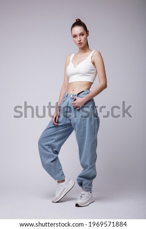High fashion photo of a beautiful elegant young woman in a pretty white top and sneakers, blue denim jeans posing over white, soft gray background. Studio Shot.