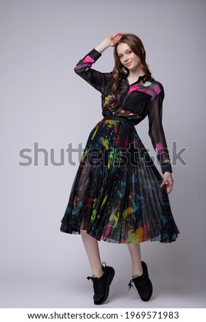 High fashion photo of a beautiful elegant young woman in a pretty black long skirt and shirt with colored patterns, sneakers posing over white, soft gray background. Studio Shot.