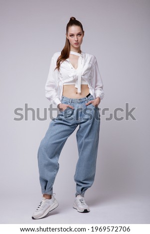 High fashion photo of a beautiful elegant young woman in a pretty white shirt and sneakers, blue denim jeans posing over white, soft gray background. Studio Shot. Royalty-Free Stock Photo #1969572706