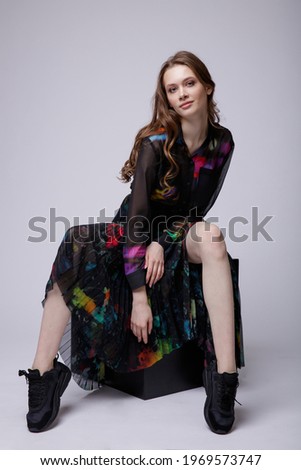 High fashion photo of a beautiful elegant young woman in a pretty black long skirt and shirt with colored patterns, sneakers posing over white, soft gray background. Studio Shot. Model sits on a cube