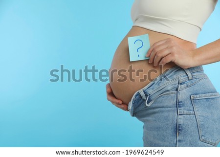 Pregnant woman with sticky note on light blue background, closeup. Choosing baby name