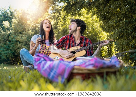 Couple of young friends singing loud together at picnic. Two young people sitting on the grass and playing guitar.