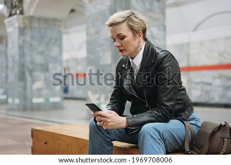 short haired caucasian woman wearing leather jacket and jeans sitting alone on  metro station reading something in smartphone waiting for train to come. Image with selective focus and noise effect