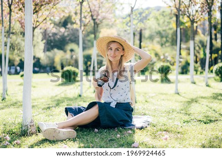 A woman traveler in a hat sits on the grass with a camera in the city garden. Travel and summer vacations.