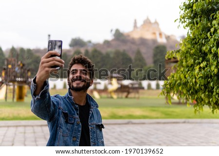 Handsome young man with curly hair and a beard wearing denim and taking a selfie with his smartphone in the pyramid of Cholula, Mexico. Royalty-Free Stock Photo #1970139652