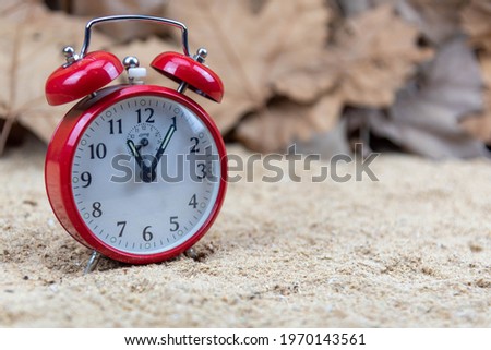 Red analog clock and maple leaves in the background. Autumn leaves and alarm clock. Vintage alarm clock. Climatic seasons. Feelings of peace and nostalgia.