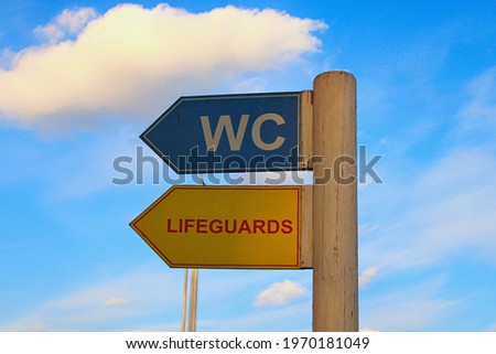 White letters ?WC? on blue arrow sign on wooden pole. Red colored word ?Lifeguard? on yellow arrow sign on wooden pole. Blue sky in the background. Venice beach in Hydropark, Kyiv, Ukraine.
