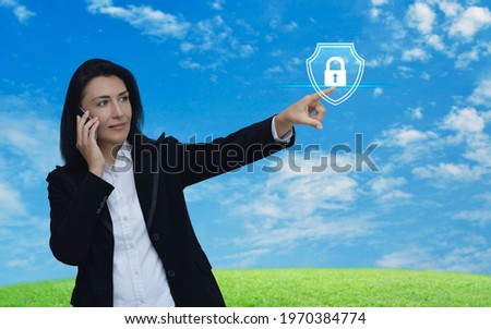 Caucasian businesswoman talking on her cell phone and pointing finger to padlock with shield icon over green grass field with blue sky, Technology security insurance online concept