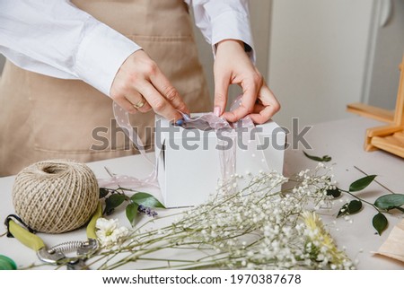A florist decorates a gift box with flowers and a ribbon on a white desktop. Only the hands are in the frame Royalty-Free Stock Photo #1970387678