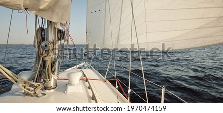 White sloop rigged yacht sailing in an open sea at sunset. Clear sky. A view from the deck to the bow, mast, sails. Transportation, travel, cruise, sport, recreation, leisure activity, racing, regatta