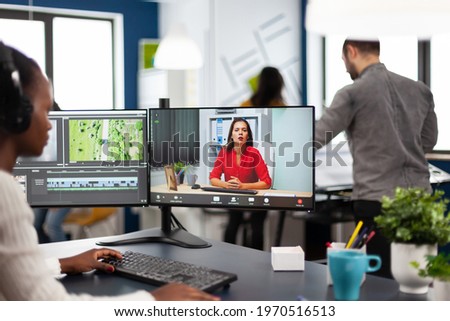 African woman editor with headset talking during online conference with partner on video call editing client work, getting feedback on commercial movie using post production software on pc in office