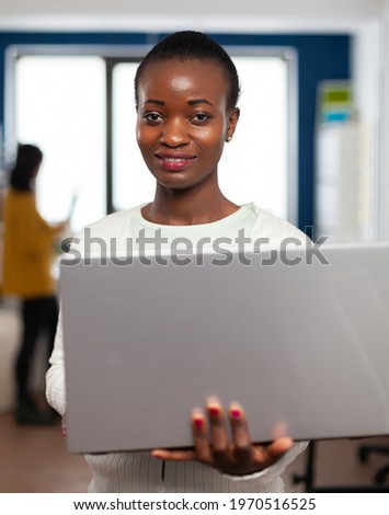 African videographer smiling at camera holding at laptop standing in creative agency office. Videographer working in multimedia studio production editing video in modern workplace.
